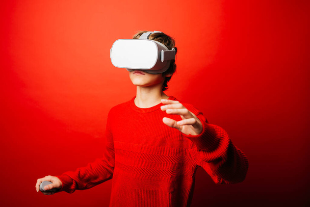 Niño preadolescente vestido de rojo sobre un fondo rojo jugando videojuegos con gafas de realidad virtual - Foto, imagen