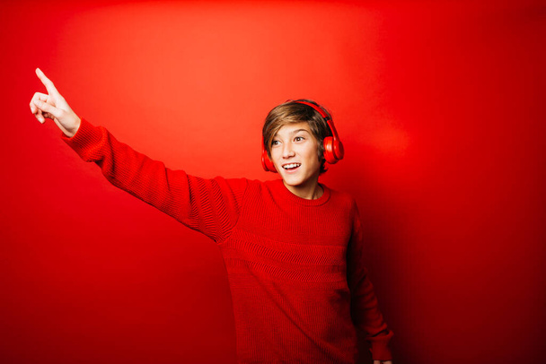 Προεφηβικό αγόρι που φοράει κόκκινο πουλόβερ και κόκκινα ακουστικά πάνω σε κόκκινο φόντο, ακούει μουσική και χορεύει - Φωτογραφία, εικόνα