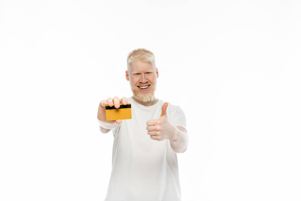 gai albinos homme en t-shirt tenant carte de crédit et montrant pouce vers le haut isolé sur blanc  - Photo, image