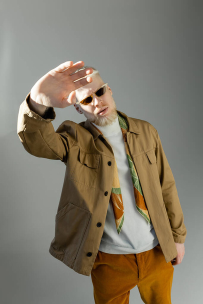 élégant homme albinos en lunettes de soleil et veste de chemise posant avec la main tendue isolé sur gris - Photo, image
