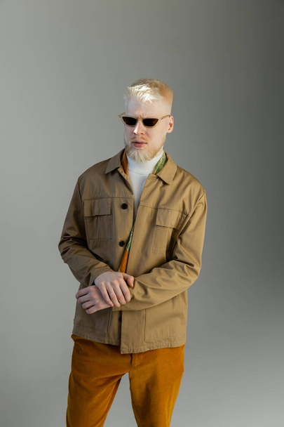 stílusos albínó férfi napszemüvegben és ing kabát áll elszigetelt szürke - Fotó, kép