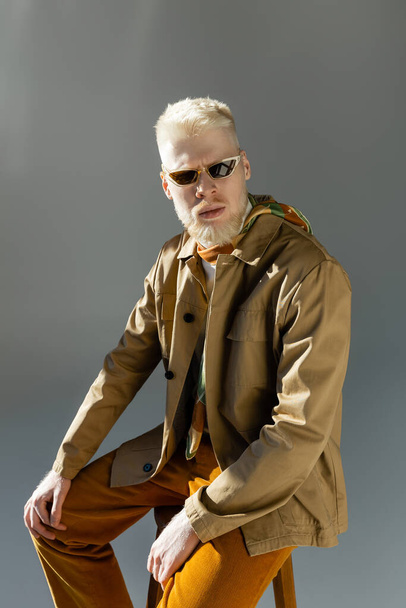 γενειοφόρος άνδρας Albino σε κομψά γυαλιά ηλίου και πουκάμισο σακάκι κάθεται σε ψηλή καρέκλα σε γκρι - Φωτογραφία, εικόνα