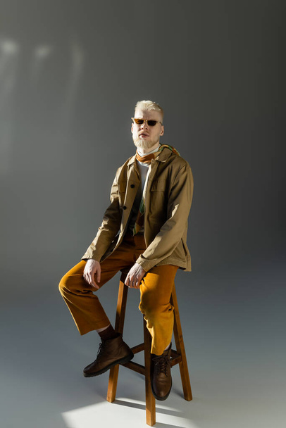 longitud completa del hombre albino barbudo en gafas de sol y chaqueta de camisa sentado en la silla alta en gris - Foto, imagen