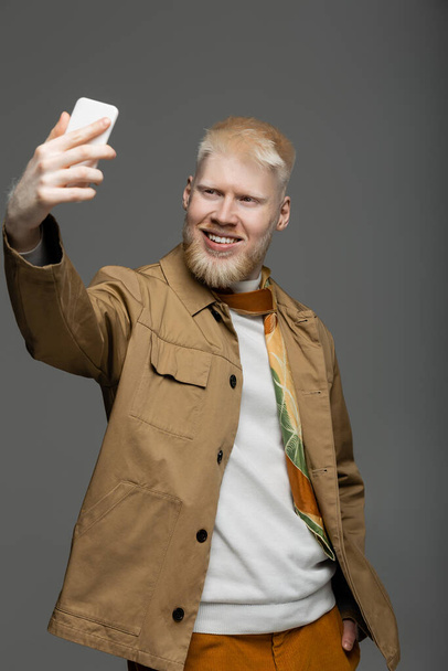 весёлый альбинос в стильной рубашке делает селфи на смартфоне, изолированном на сером  - Фото, изображение