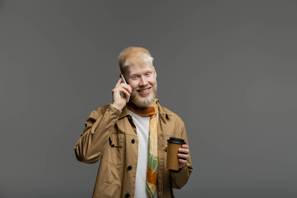 happy albino man in shirt jacket holding paper cup and talking on smartphone isolated on grey - Photo, Image