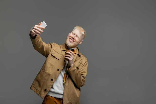 довольный альбинос мужчина в рубашке куртка держа бумажную чашку и делая селфи на смартфоне изолированы на серый - Фото, изображение