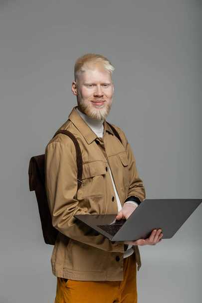 χαρούμενο αλμπίνο φοιτητής με σακίδιο πλάτης χρησιμοποιώντας φορητό υπολογιστή, ενώ η μελέτη σε απευθείας σύνδεση απομονωμένη σε γκρι - Φωτογραφία, εικόνα