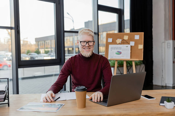 vrolijke albino zakenman in bril op zoek naar laptop in de buurt van papieren beker en infographics op het bureau   - Foto, afbeelding