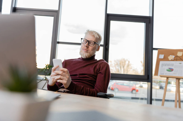 homme d'affaires barbu albinos dans des lunettes en utilisant un smartphone près d'un ordinateur portable dans le bureau  - Photo, image