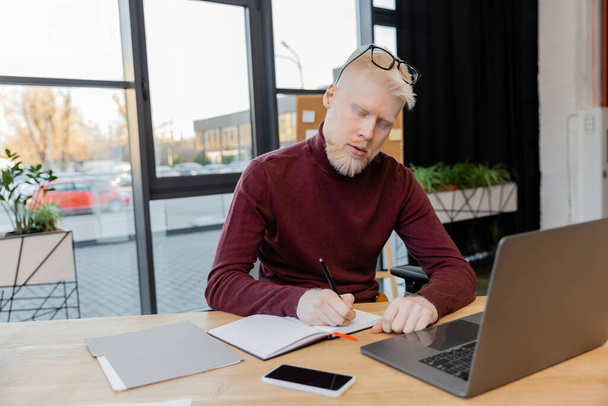 Bärtiger Albino-Geschäftsmann mit Brille schreibt auf Notizbuch neben Gadgets auf Schreibtisch  - Foto, Bild