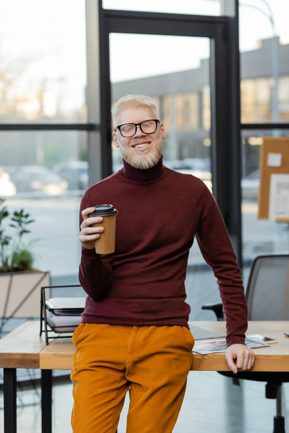 bearded albino businessman in glasses smiling and holding coffee to go in office - Photo, Image