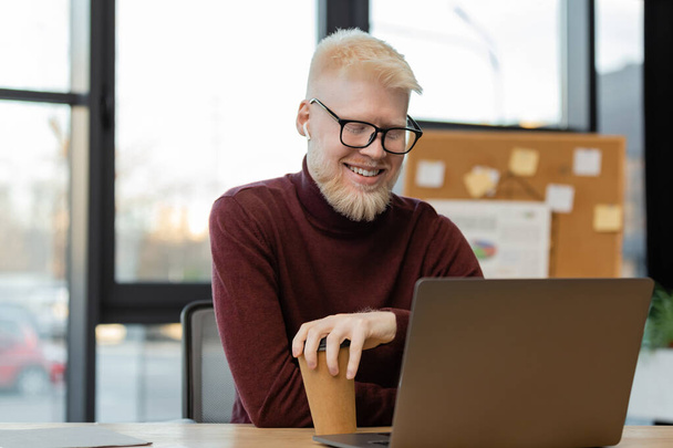 empresário albino alegre em óculos olhando para laptop e segurando copo de papel no escritório  - Foto, Imagem