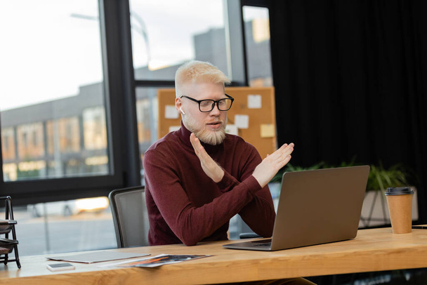 bebaarde albino zakenman in oortelefoons tonen afval gebaar tijdens videogesprek op laptop tijdens het werk in het kantoor  - Foto, afbeelding