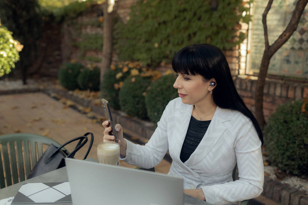 屋外カフェに座っている幸せな中年女性のショット。オンラインで働く間、カジュアルな服を着てラップトップを使用している自信のある実業家。オンライン作業概念 - 写真・画像