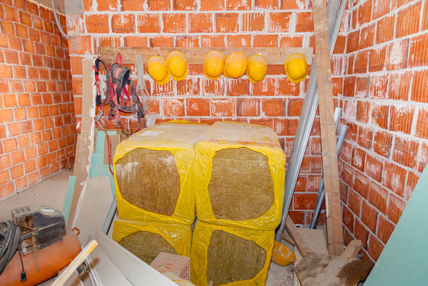 Package of thermal insulation material, rock wool and workers' industrial work equipment, yellow helmets and safety belts hang on a makeshift hanger made of wooden board and nails placed in warehouse. - Photo, Image