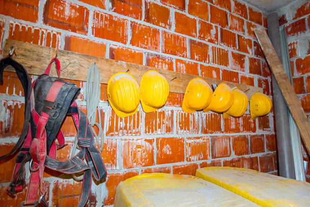 Pracovnické pracovní zařízení, žluté přilby a bezpečnostní pásy visí na provizorním ramínku z dřevěné desky a hřebíků, připevněném na zdi z červených cihel. - Fotografie, Obrázek