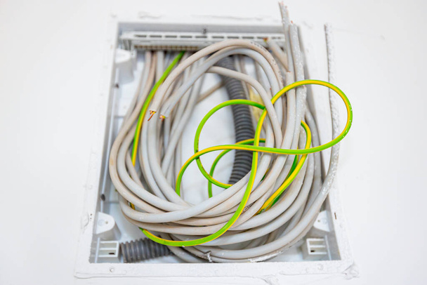 Bundle of various cables hang from a plastic fuse box mounted on the plaster wall, building under construction, work in progress. Installation of electrical wires and cables. - Photo, Image