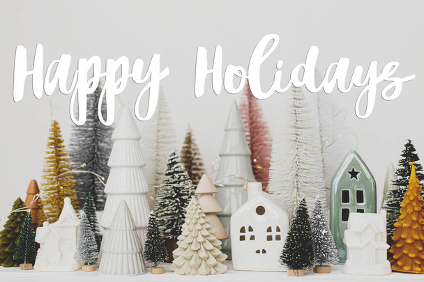Happy Holidays znak tekstowy na stylowe choinki i dekoracje domów na białym stole. Nowoczesna scena świąteczna, miniaturowa, przytulna, śnieżna wioska. Kartka z życzeniami sezonu - Zdjęcie, obraz