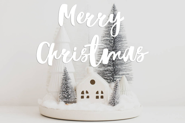 Frohe Weihnachten Text auf stilvollen kleinen Weihnachtsbäumen und Haus auf weißem Tisch. Die Grußkarte der Saison. Gemütliche Weihnachtszauberszene, verschneites Dorf im Miniaturformat. Handschriftliches Zeichen - Foto, Bild