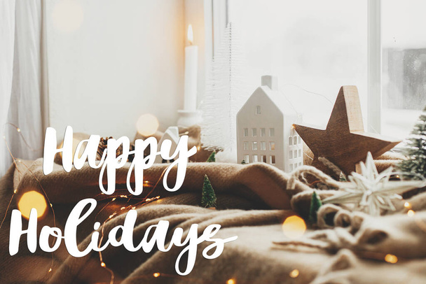 Happy Holidays tekstbord op stijlvolle kerstversiering, boom, verlichting, huisje en houten ster op gezellige deken bij besneeuwd raam. Groetkaart van het seizoen - Foto, afbeelding