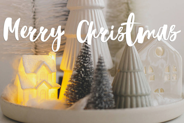 Καλά Χριστούγεννα κείμενο για τα κομψά μικρά χριστουγεννιάτικα δέντρα και το σπίτι σε λευκό τραπέζι. Ευχετήρια κάρτα εποχής. Άνετο Χριστούγεννα μαγική σκηνή, μινιατούρα χιονισμένο χωριό με φώτα. Χειρόγραφο σήμα - Φωτογραφία, εικόνα