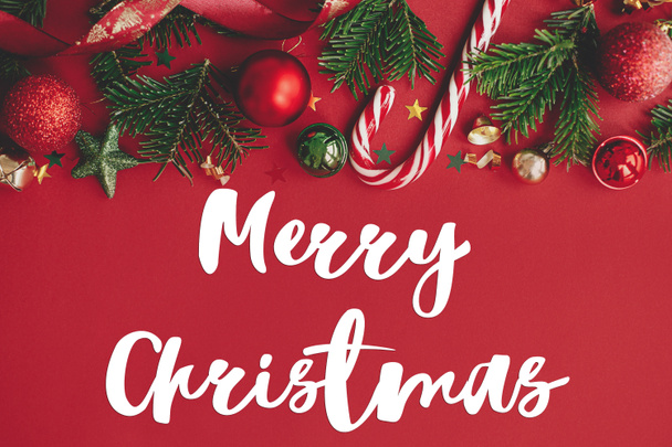 Καλά Χριστούγεννα κείμενο σε στολίδια Χριστούγεννα, κλαδιά ελάτης, κομφετί και ζαχαροκάλαμο καραμέλα σε κόκκινο φόντο επίπεδη θέσει. Ευχετήρια κάρτα εποχής. Χειρόγραφο σήμα - Φωτογραφία, εικόνα