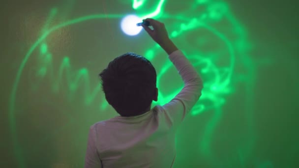 fysica en ontwikkeling van kinderen, nieuwsgierige jongen met zaklamp in handen tekent met licht op groene muur in een interactieve ruimte - Video