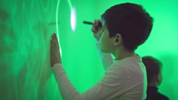 moderne opvoeding van kinderen, geïnteresseerde jongen tekent patroon met een ultraviolet zaklamp op een groene muur in een interactieve ruimte - Video