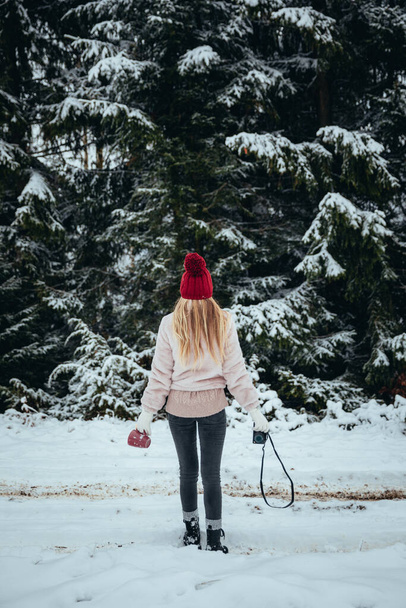 バックビューのブロンド女性とともにカメラと赤いカップで手に立って雪に覆われた森   - 写真・画像