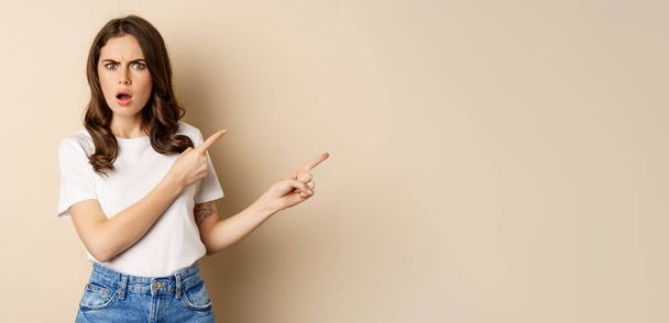 Frustrierte Frau sieht beleidigt aus, zeigt mit dem Finger nach rechts und keucht geschockt, steht im T-Shirt vor beigem Hintergrund. - Foto, Bild