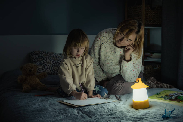 Mãe e filhinha estudando e desenhando em uma escuridão completa durante a falta de eletricidade. Menina usa lanterna de acampamento para fazer seu dever de casa durante o apagão. Conceito de crise energética - Foto, Imagem