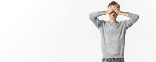 Image of excited middle-aged guy in grey sweater, cover eyes with hands and looking amazed while waiting for surprise, playing hide-n-seek, standing over white background. - Photo, Image