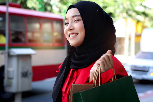 Szczęśliwy uśmiechnięty młody azjatycki muzułmanin kobiety noszenie czarny chusta hidżab w czerwonej sukience trzymając torby na zakupy na ulicy spaceru, portret atrakcyjnej pięknej dziewczyny z torby na zakupy spacery po drodze miasta - Zdjęcie, obraz