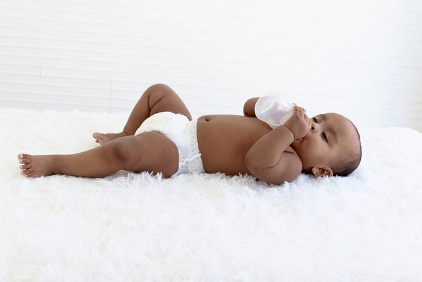 Adorabile un bambino afro-americano strisciante di sei mesi sdraiato su un soffice tappeto bianco, felice dolce bambino bambina che tiene in mano la bottiglia di latte e mangia cibo, concetto di cura del bambino di alimentazione infantile - Foto, immagini
