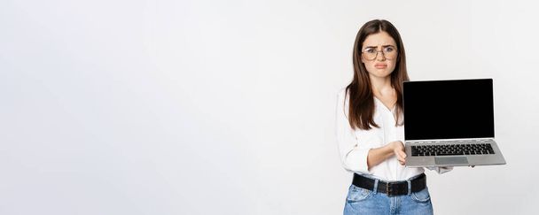Sad brunette girl, student showing laptop screen and grimacing upset, standing disappointed against white background. - Fotografie, Obrázek