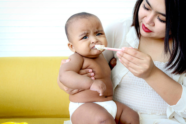 Madre alimentando a su linda niña Asiática Afroamericana bebé hija con cuchara de comida, bebé niño disfrutar de comer sano casero bebé comida para mamá en casa. - Foto, imagen
