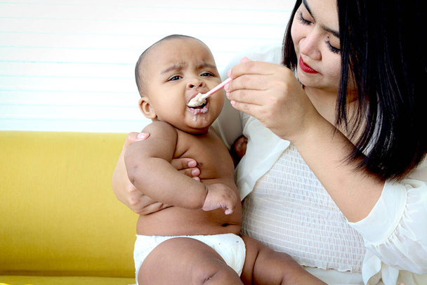 Madre che alimenta la sua bambina carina Figlia afroamericana asiatica con cucchiaio di cibo, il bambino bebè si diverte a mangiare cibo per bambini sano fatto in casa per la mamma a casa. - Foto, immagini
