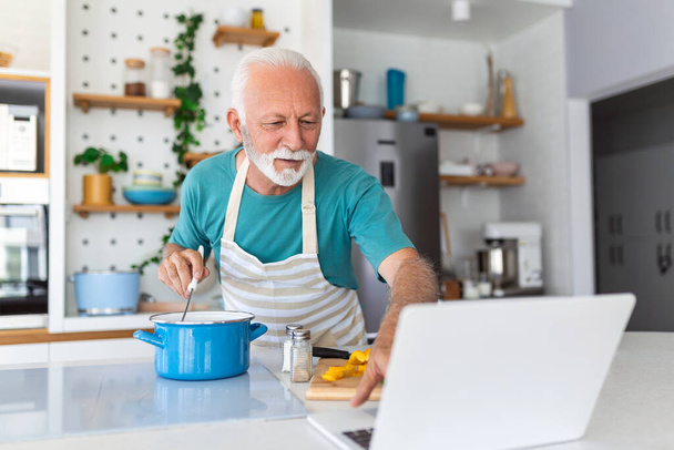 Счастливый пожилой человек с удовольствием готовит дома - Пожилой человек готовит здоровый обед на современной кухне, глядя на приемник на своем ноутбуке - Фото, изображение