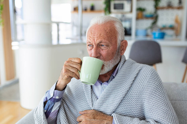 Starszy mężczyzna cierpiący na grypę pijący herbatę siedząc owinięty w koc na kanapie w domu. Chorym starszym mężczyzną z bólem głowy siedzącym pod kocem w salonie. Człowiek z przeziębieniem leżący na kanapie trzymając kubek gorącej herbaty. - Zdjęcie, obraz