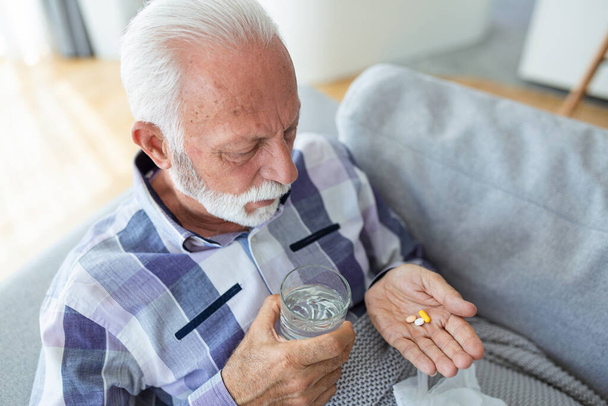 Senior man taking pills while sitting on couch at home, holding white jar with treatment, copy space. Grey-haired elderly man using supplements or vitamins. Healthy lifestyle in senior age concept - Photo, Image