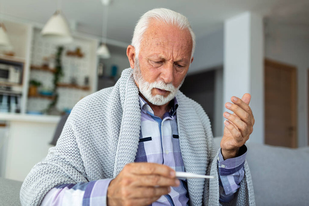 Chorzy starsi mężczyźni sprawdzają temperaturę, cierpiący na grypę sezonową lub przeziębienie leżące na kanapie, cierpiący na grypę sezonową lub przeziębienie. - Tak. Seniorzy czują się niezdrowi z grypą w domu. - Zdjęcie, obraz