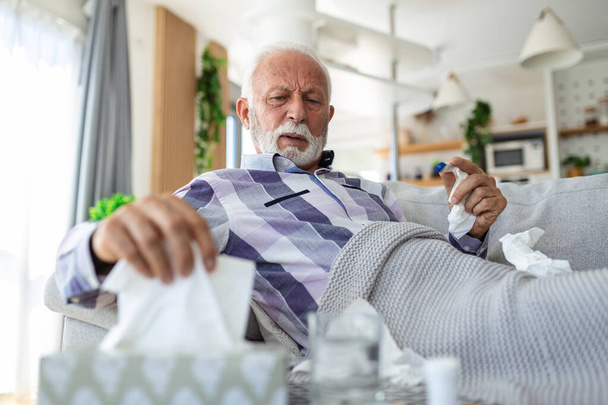 Kranker älterer Mann, der seine Temperatur überprüft und an saisonaler Grippe oder Erkältung leidet, liegt auf dem Sofa und leidet unter saisonaler Grippe oder Erkältung. . Kranke Senioren fühlen sich bei Grippe zu Hause ungesund - Foto, Bild