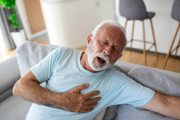 Starszy mężczyzna przyciska rękę do klatki piersiowej ma atak serca cierpi na nieznośny ból, Dojrzały mężczyzna z bólem serca w salonie. Starszy mężczyzna cierpiący na silny ból w klatce piersiowej atak serca w domu - Zdjęcie, obraz