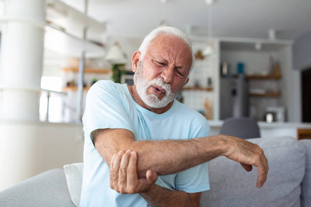 Kolu ağrıyan yaşlı bir adam. İçeride acı çeken yaşlı bir erkek el masajı yapıyor. Dirseğini tutan yaşlı adam dirsek ağrısından muzdarip. Evdeki acıdan muzdarip son sınıf öğrencisi. Yaşlılık, sağlık - Fotoğraf, Görsel