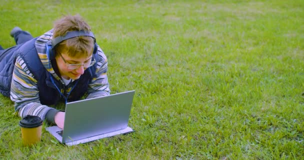 Kulaklık takan bir genç diz üstü bilgisayar monitörünün önünde çimlere uzanır. Ödevini yapar ya da sosyal ağlardan arkadaşlarıyla internetten iletişim kurar. 4k görüntü. - Video, Çekim