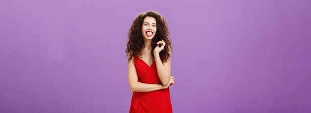 Studio shot of playful and joyful artistic caucasian female. student in red evening dress sticking out tongue happily having fun playing with curl standing over purple background careless and carefree - Photo, Image