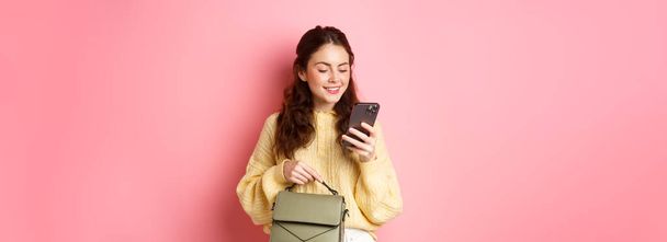Glamour dívka drží kabelku a textové zprávy na smartphonu, čtení obrazovky mobilního telefonu s bezstarostným úsměvem, stojící nad růžovým pozadím. - Fotografie, Obrázek