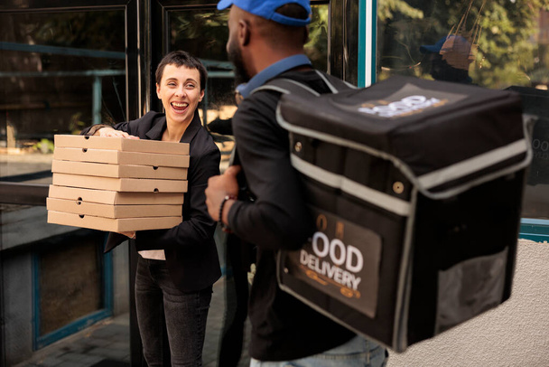 Cliente feliz sosteniendo la pila de cajas de pizza, recibiendo orden de mensajero al aire libre, mujer tomando paquetes de comida pila para colegas. Hombre afroamericano entregando almuerzo de oficina, servicio de entrega de comida - Foto, imagen