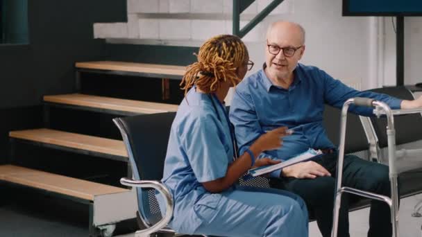 Medische assistent adviserende oudere man met loopframe, zittend in het ziekenhuis receptie lobby. Verpleegster in wachtkamer in gezondheidscentrum in gesprek met oude man, invullen van klinische check-up rapport. - Video