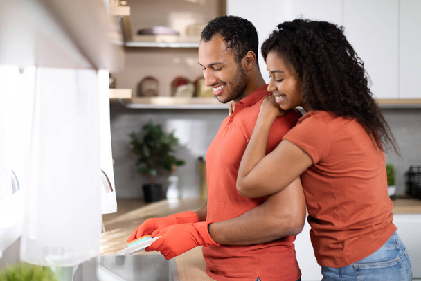Домашние хлопоты по дому. Рад, что чернокожая жена обнимает мужа в резиновых перчатках, парень моет посуду в кухонном интерьере, копирует пространство. Любовь, романтика, семейные отношения и образ жизни - Фото, изображение